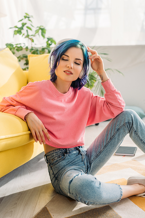 Woman with colorful hair, closed eyes and headphones listening music near sofa on floor