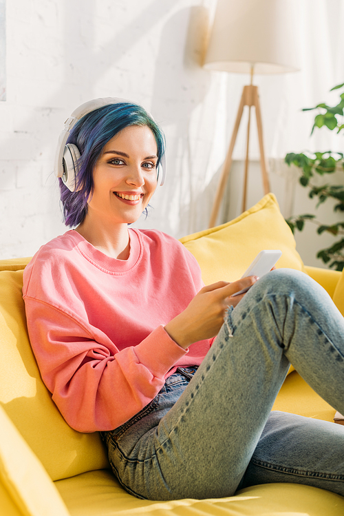 Woman with colorful hair and headphones holding smartphone, smiling and  on sofa