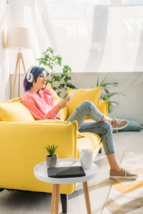 Woman with colorful hair and headphones holding smartphone on sofa near coffee table in living room