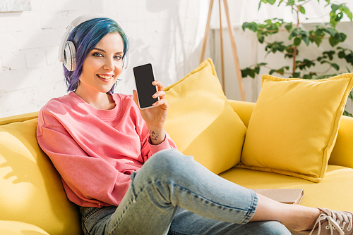 Woman with colorful hair and headphones smiling,  and showing smartphone on sofa in living room