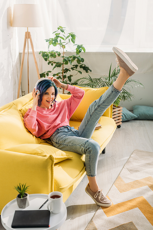 Woman with colorful hair having fun with headphones and raised leg on sofa in living room