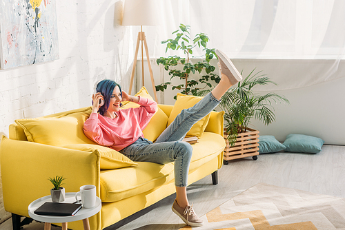 Woman with colorful hair, headphones and raised leg smiling on sofa in living room