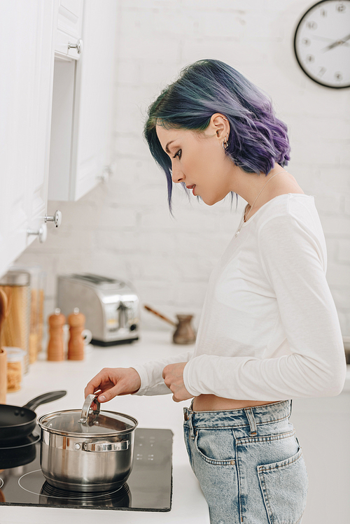 Girl with colorful hair preparing food and touching pan lid near kitchen stove