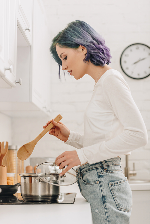 Girl with colorful hair holding pan lid and spatula near kitchen stove