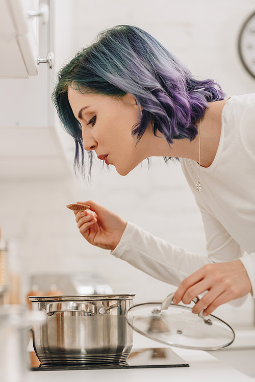 Selective focus of girl with colorful hair holding pan lid and blowing on spatula near kitchen stove