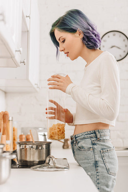 Selective focus of girl with colorful hair holding container with pasta near kitchen stove