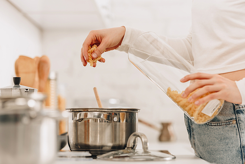 Cropped view of girl putting pasta in pan near kitchen stove