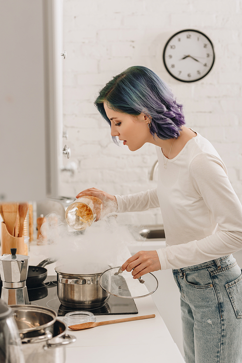 Selective focus of girl with colorful hair putting pasta in pan with steam near kitchen stove