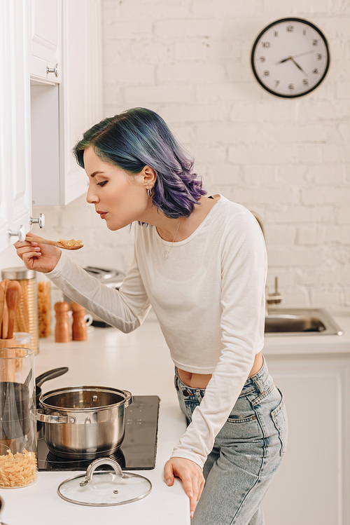 Girl with colorful hair blowing on spatula with pasta near kitchen stove