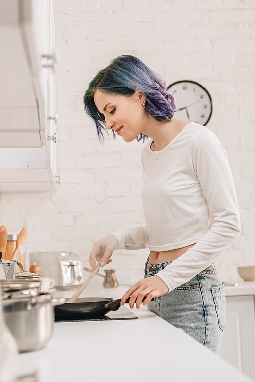 Selective focus of girl with colorful hair preparing food with spatula on frying pan in kitchen