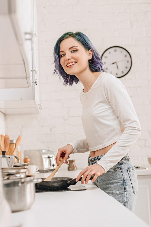 Low angle view of girl with colorful hair preparing food with spatula on frying pan, smiling and  in kitchen