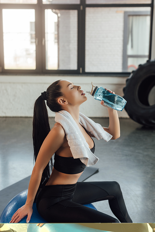 Beautiful sportswoman with closed eyes and towel drinking water and sitting on fitness ball in gym