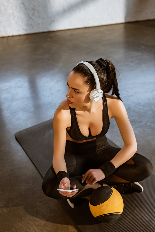 High angle view of sportswoman with crossed legs in headphones holding smartphone near ball on fitness mat in gym