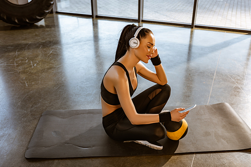 Sportswoman with crossed legs in headphones using smartphone and smiling near ball on fitness mat in gym