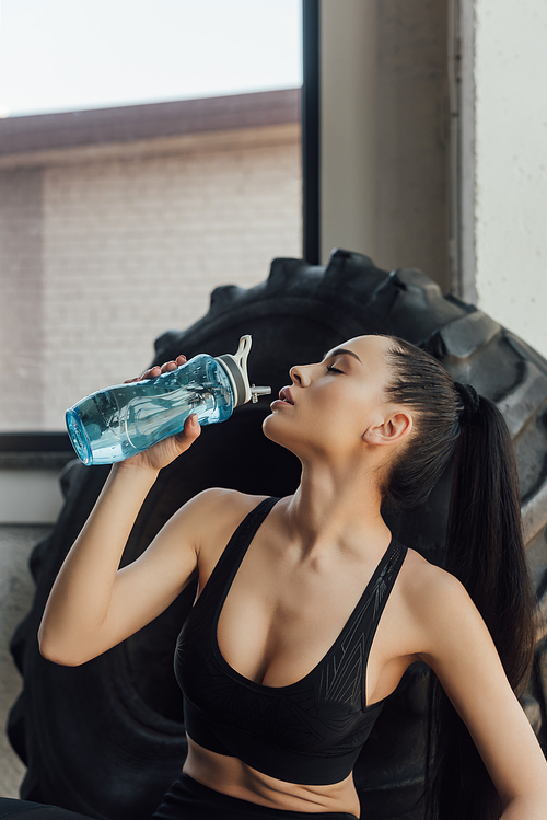 Sexy sportswoman with closed eyes drinking water near tire in gym