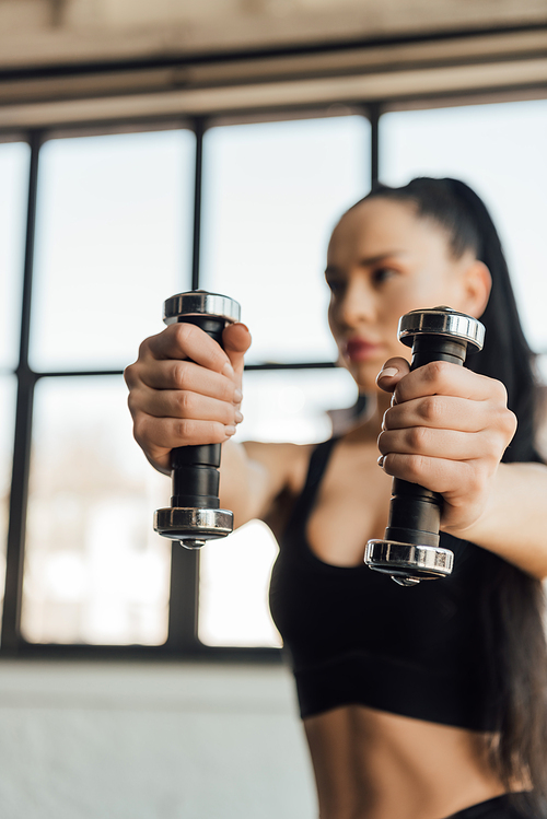 Selective focus of sportswoman holding dumbbells with outstretched hands in gym