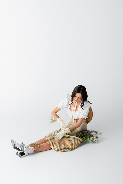 young woman in spring outfit and gloves sitting near flowers in straw bag on white