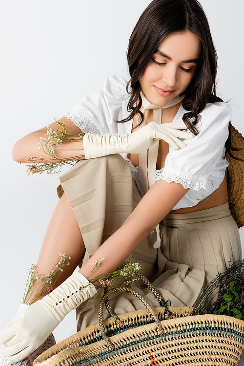 smiling woman in spring outfit and gloves with flowers sitting while posing isolated on white
