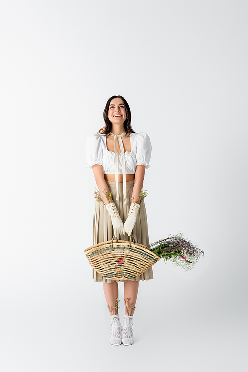 full length of happy young woman in spring outfit holding straw bag with flowers on white
