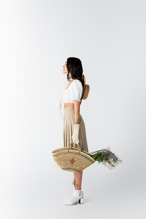 side view of young woman in spring outfit holding straw bag with flowers on white