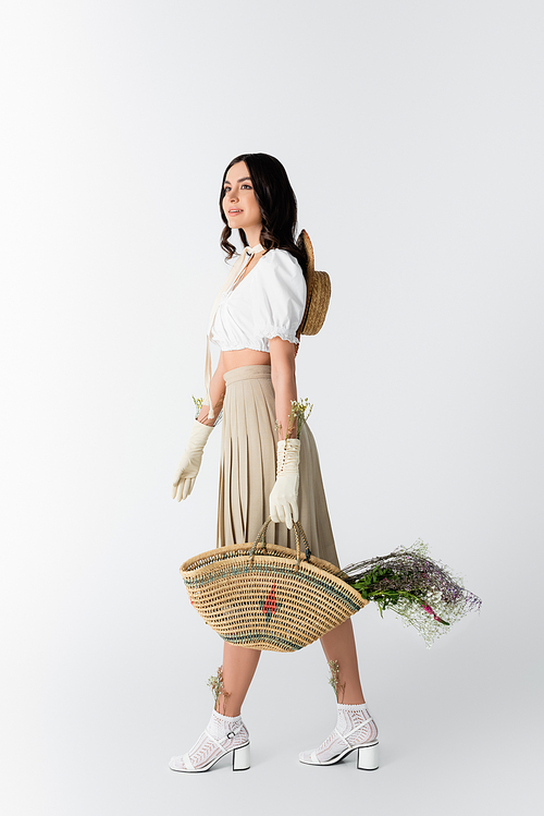 full length of happy woman in spring outfit holding straw bag with flowers on white