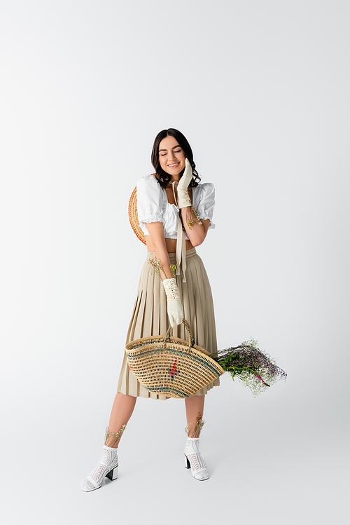 full length of smiling young woman in spring outfit holding straw bag with flowers on white