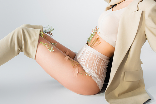 cropped view of model in blazer, crop top and panties with blooming flowers sitting while posing on white
