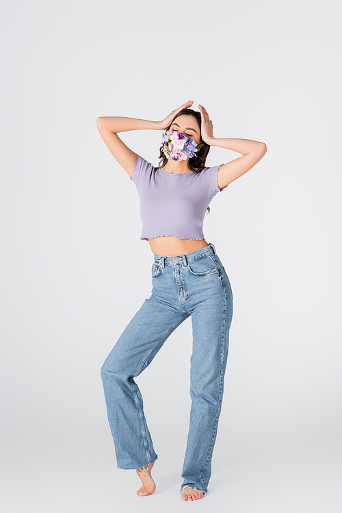 full length of young woman in crop top, jeans and medical mask with blooming flowers posing on white
