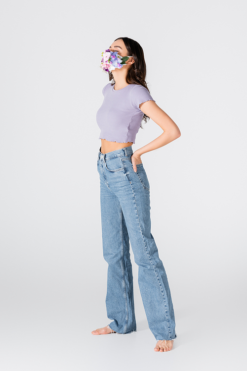 full length of young model in crop top, jeans and medical mask with blooming flowers posing with hand in pocket on white
