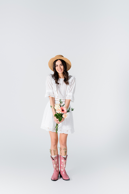 full length of smiling young woman in straw hat and dress holding bouquet of spring flowers on white