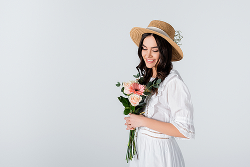 cheerful young woman in straw hat and dress holding bouquet of spring flowers isolated on white