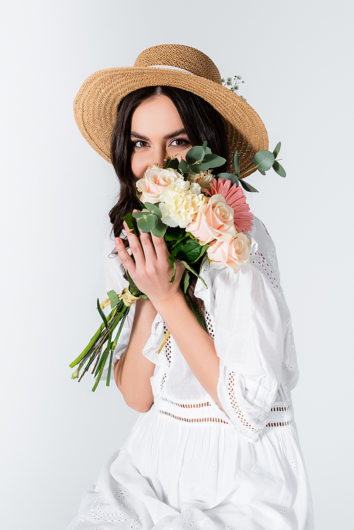 young woman in straw hat and dress covering face with bouquet of spring flowers isolated on white