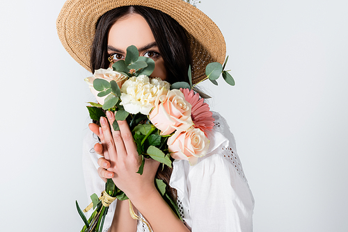 young model in straw hat and dress covering face with bouquet of spring flowers isolated on white