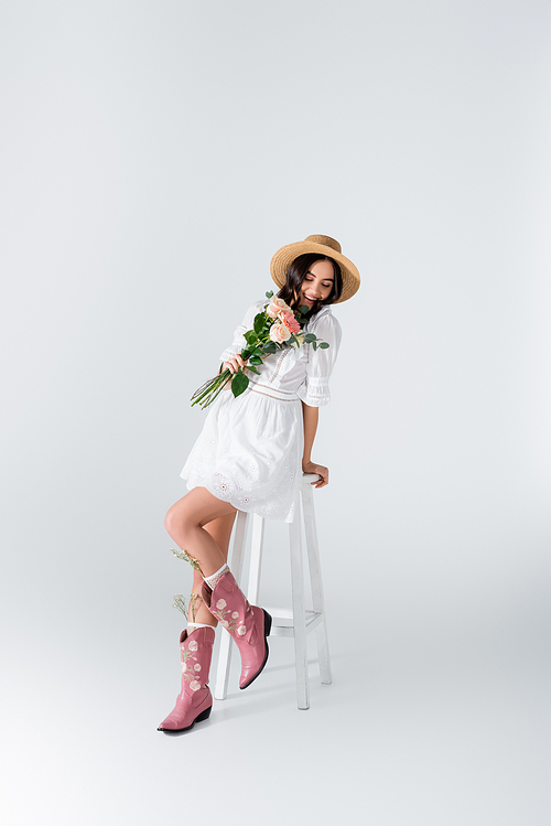 joyful woman in straw hat and dress sitting on chair and holding bouquet of spring flowers on white