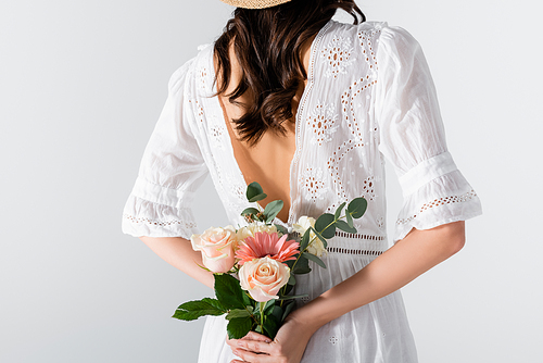 cropped view of young woman in dress holding bouquet of spring flowers behind back isolated on white