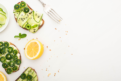 top view of fresh cucumber toasts with seeds, mint leaves near lemon and fork on white background