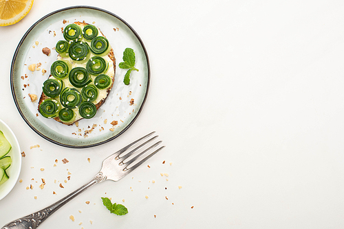 top view of fresh cucumber toast with sesame and mint leaves on plate near fork and lemon on white background
