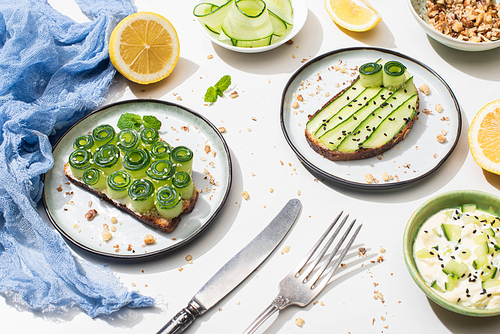 fresh cucumber toasts on plates near cutlery, yogurt and lemon on white background