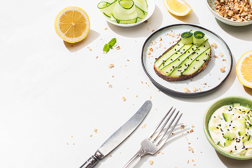 fresh cucumber toast with seeds near mint leaves, cutlery, yogurt and lemon on white background