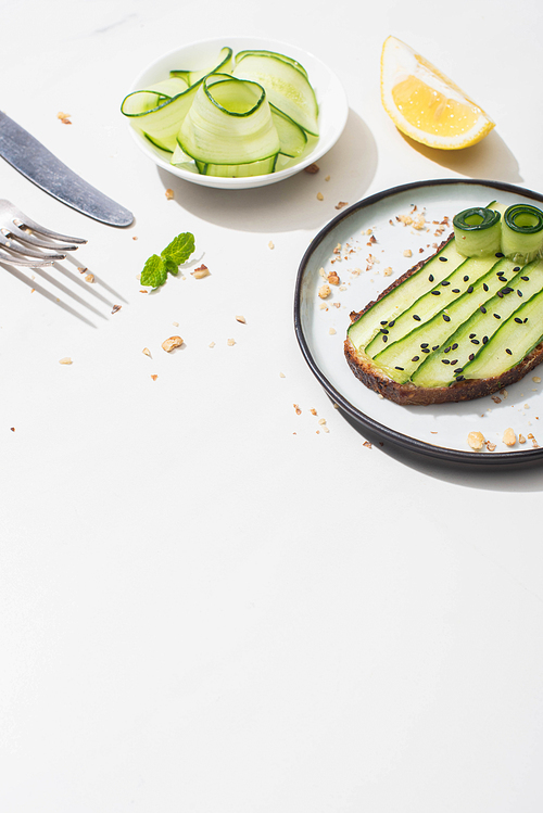 fresh cucumber toast with seeds near mint leaves, cutlery and lemon on white background