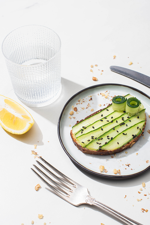 fresh cucumber toast with seeds served with cutlery, lemon and water on white background