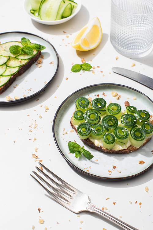 fresh cucumber toasts with seeds, mint leaves and lemon near glass of water and cutlery on white background