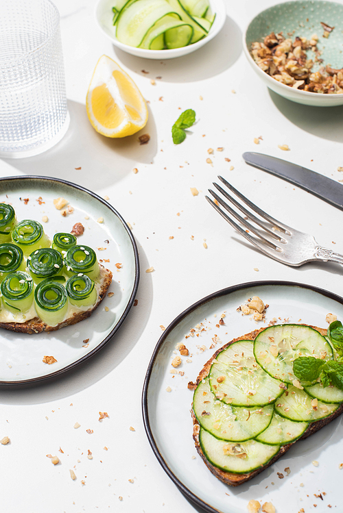 fresh cucumber toasts with seeds, mint leaves and lemon near glass of water and cutlery on white background