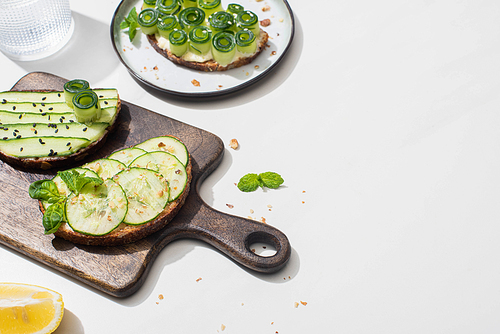 fresh cucumber toasts on wooden cutting board near water and lemon on white background