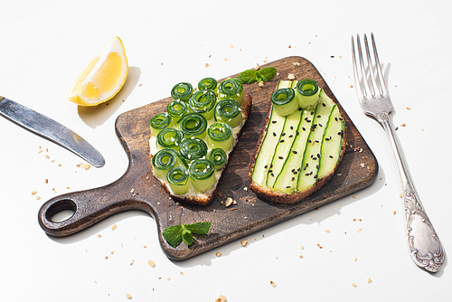 fresh cucumber toasts on wooden cutting board near cutlery and lemon on white background