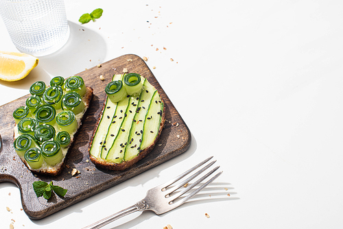 fresh cucumber toasts on wooden cutting board near water, fork and lemon on white background