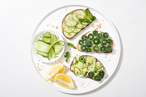 top view of fresh cucumber toasts with seeds, mint and basil leaves on plate with lemon on white background