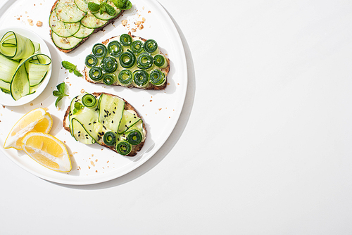top view of fresh cucumber toasts with seeds, mint and basil leaves on plate with lemon on white background