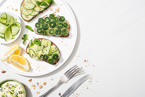 top view of fresh cucumber toasts with seeds, mint and basil leaves on plate with lemon near cutlery and yogurt on white background