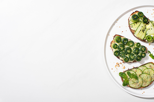 top view of fresh cucumber toasts with seeds, mint and basil leaves on plate on white background